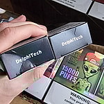 A person holding two boxes labeled "DelphiTech" next to a carton containing products with "15000 PUFFS" branding and a warning label about nicotine.