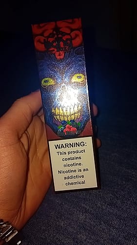 A hand holding a colorful vape box with a warning label stating, "This product contains nicotine. Nicotine is an addictive chemical."