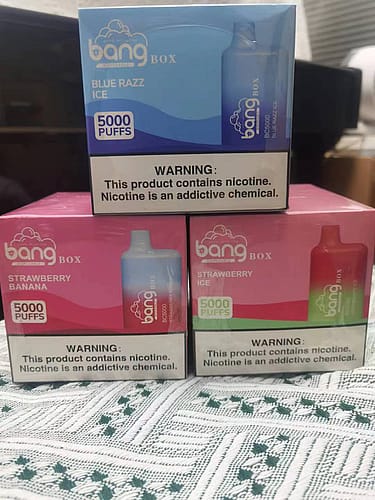 Three boxes of Bang disposable vapes in different flavors (Blue Razz Ice, Strawberry Banana, and Strawberry Ice) stacked on a patterned surface. Each box is labeled with "5000 PUFFS" and has a nicotine warning at the bottom.