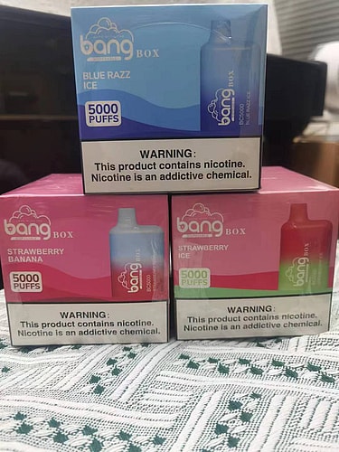 Three boxes of Bang disposable vapes in different flavors (Blue Razz Ice, Strawberry Banana, and Strawberry Ice) stacked on a patterned surface. Each box is labeled with "5000 PUFFS" and has a nicotine warning at the bottom.