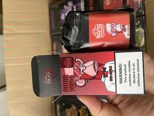 A hand holding a black and red vape device box labeled "Bang 8000 Puffs" with a cartoon ape and the flavor "Red Apple." A nicotine warning is visible on the box.