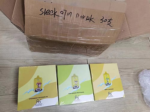 A cardboard box with black handwritten text on top, containing three packaged boxes labeled "STRAWBERRY WATERMELON," "KIWI PASSION FRUIT GUAVA," and "BERRY LEMONADE," each with a yellow and white design.