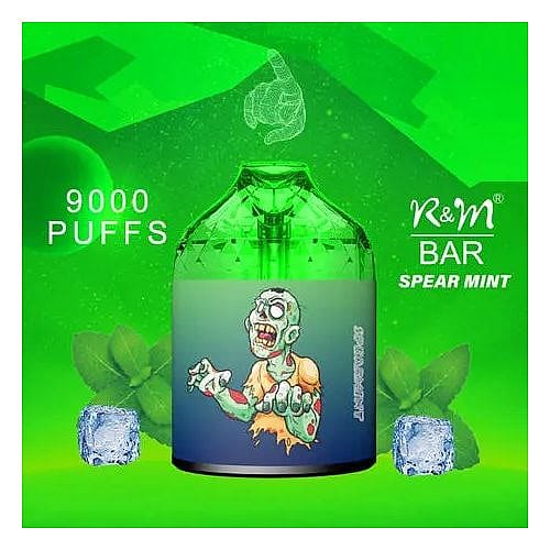 Un dispositivo per svapare verde con l'immagine di uno zombie in stile cartone animato, etichettato "9000 PUFFS" e "R&M BAR SPEAR MINT" con foglie di menta e cubetti di ghiaccio sullo sfondo.