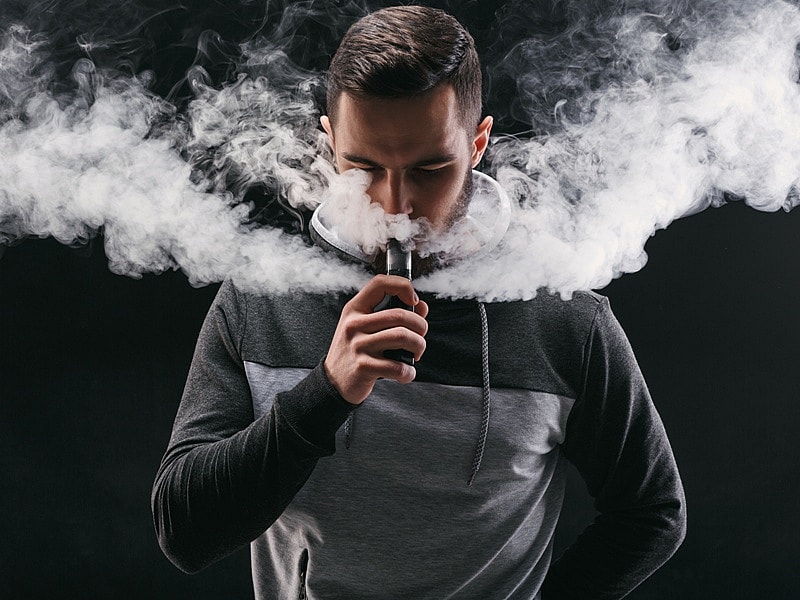 Un homme en sweat à capuche vapote, entouré d'épais nuages de vapeur.