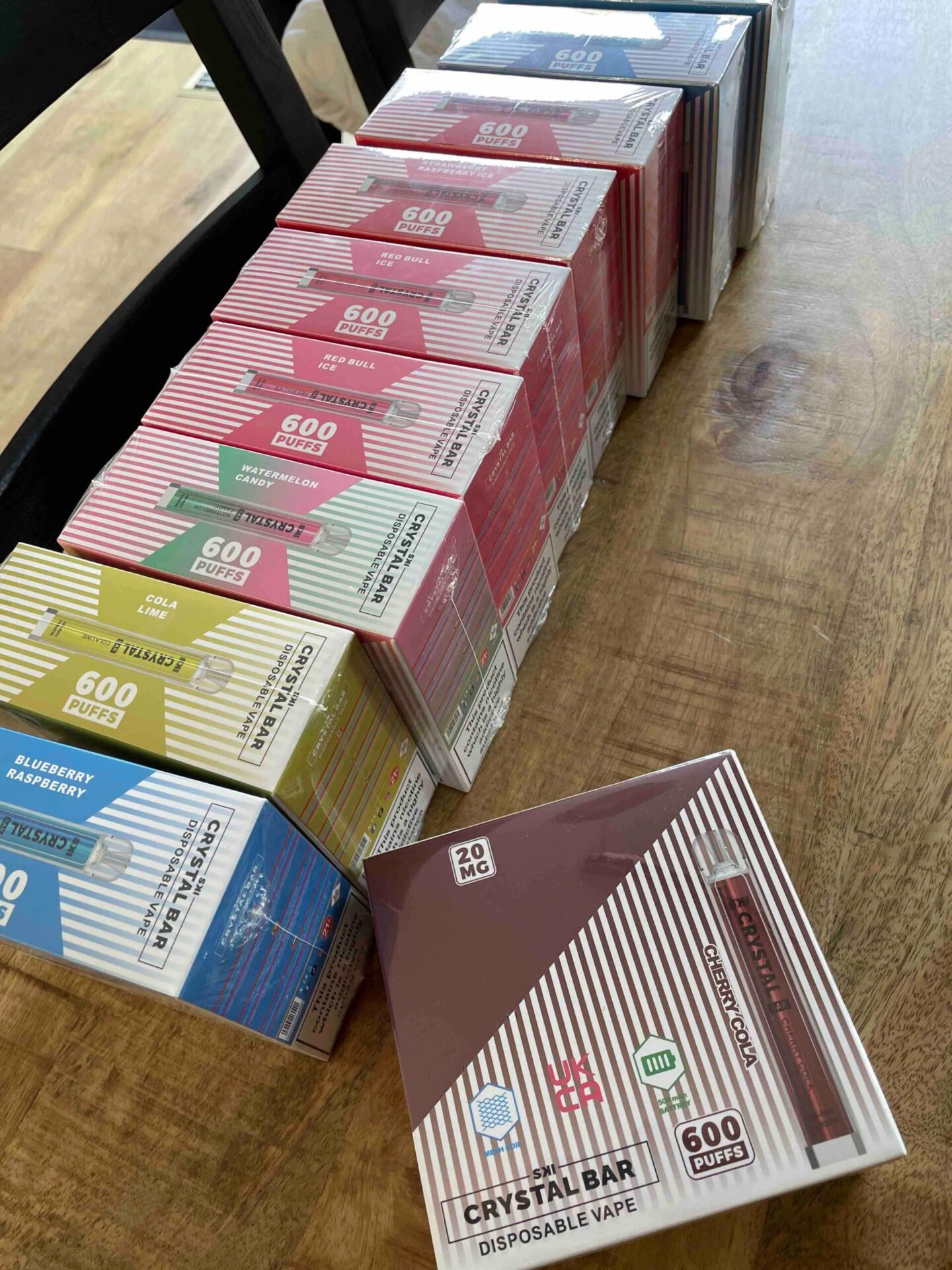 Boxes of Crystal Bar disposable vapes in various flavors, including Cola Lime, Blueberry Raspberry, Watermelon Candy, and Cherry Cola, arranged on a wooden table.