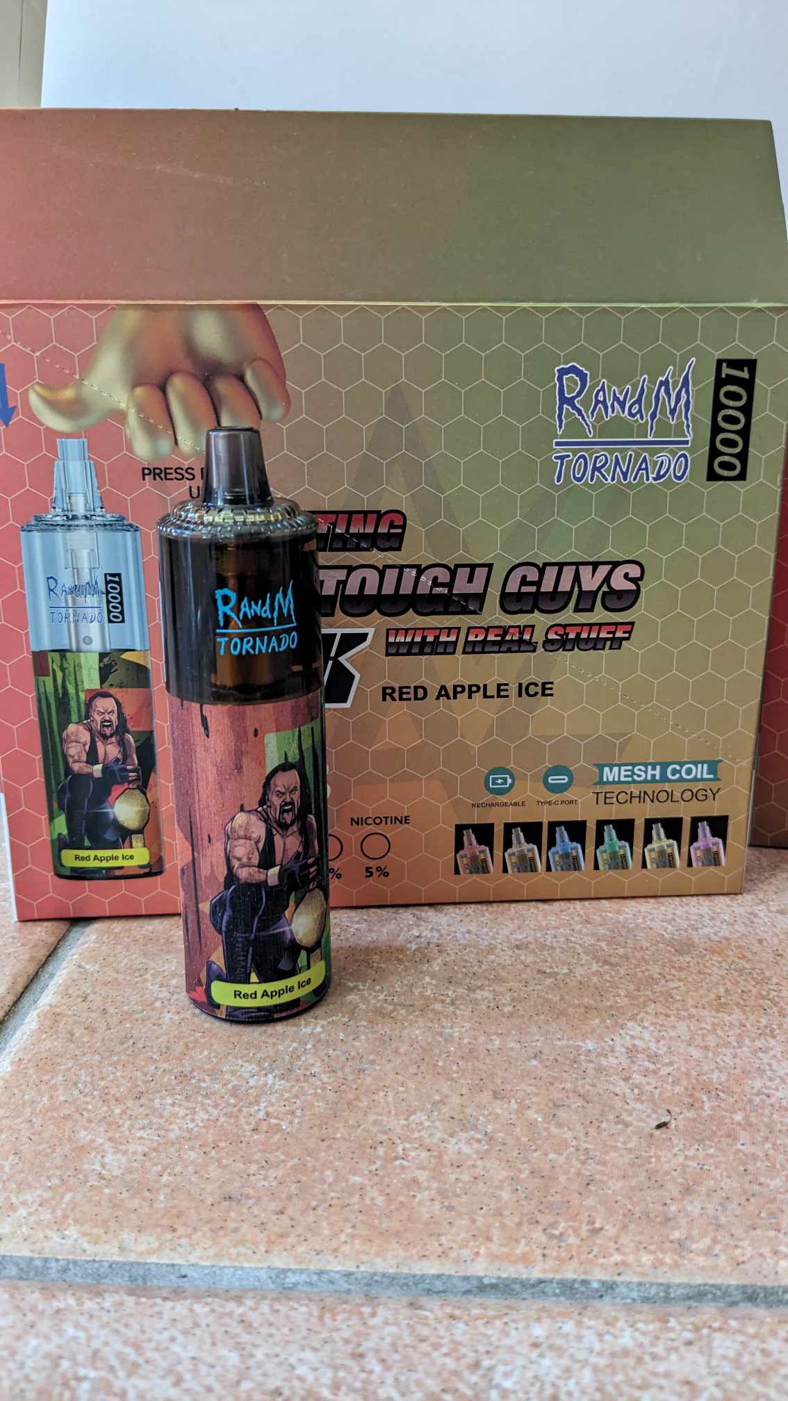 RandM Tornado vape device and packaging featuring a "Red Apple Ice" flavor. The packaging showcases a muscular cartoon character holding an apple and includes details such as "10000 puffs" and "Mesh Coil Technology."