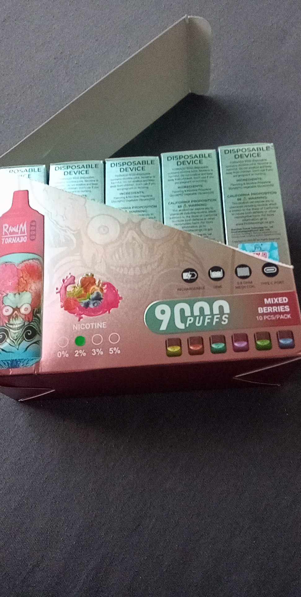 Box of disposable vape devices labeled "R&M Tornado" with 9000 puffs, mixed berries flavor, and multiple nicotine levels, showing 10 individually packed devices.