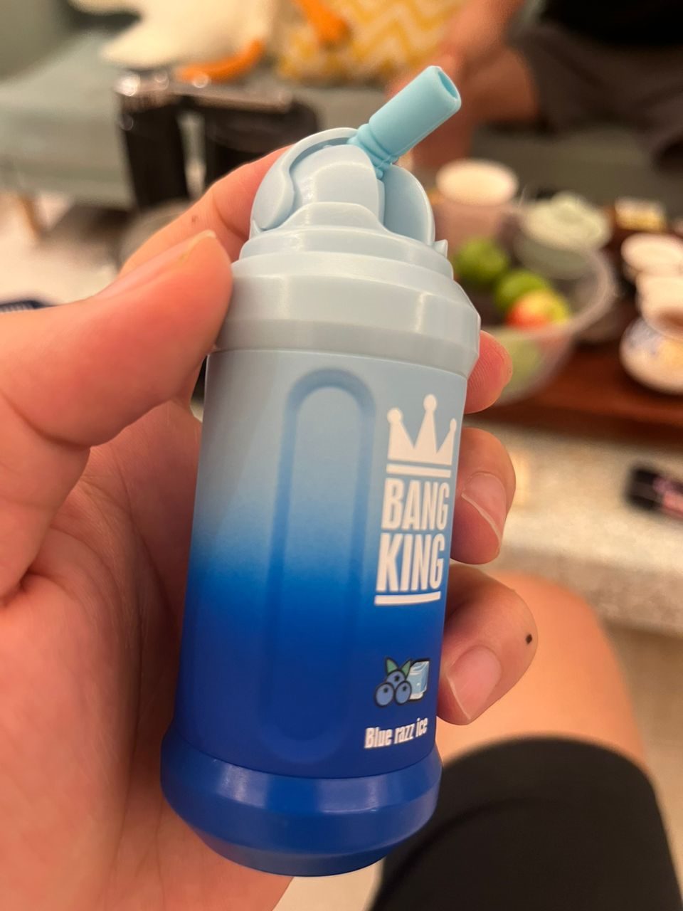 A hand holding a blue and white cylindrical object labeled "Bang King" with a small straw at the top and a blurry background.