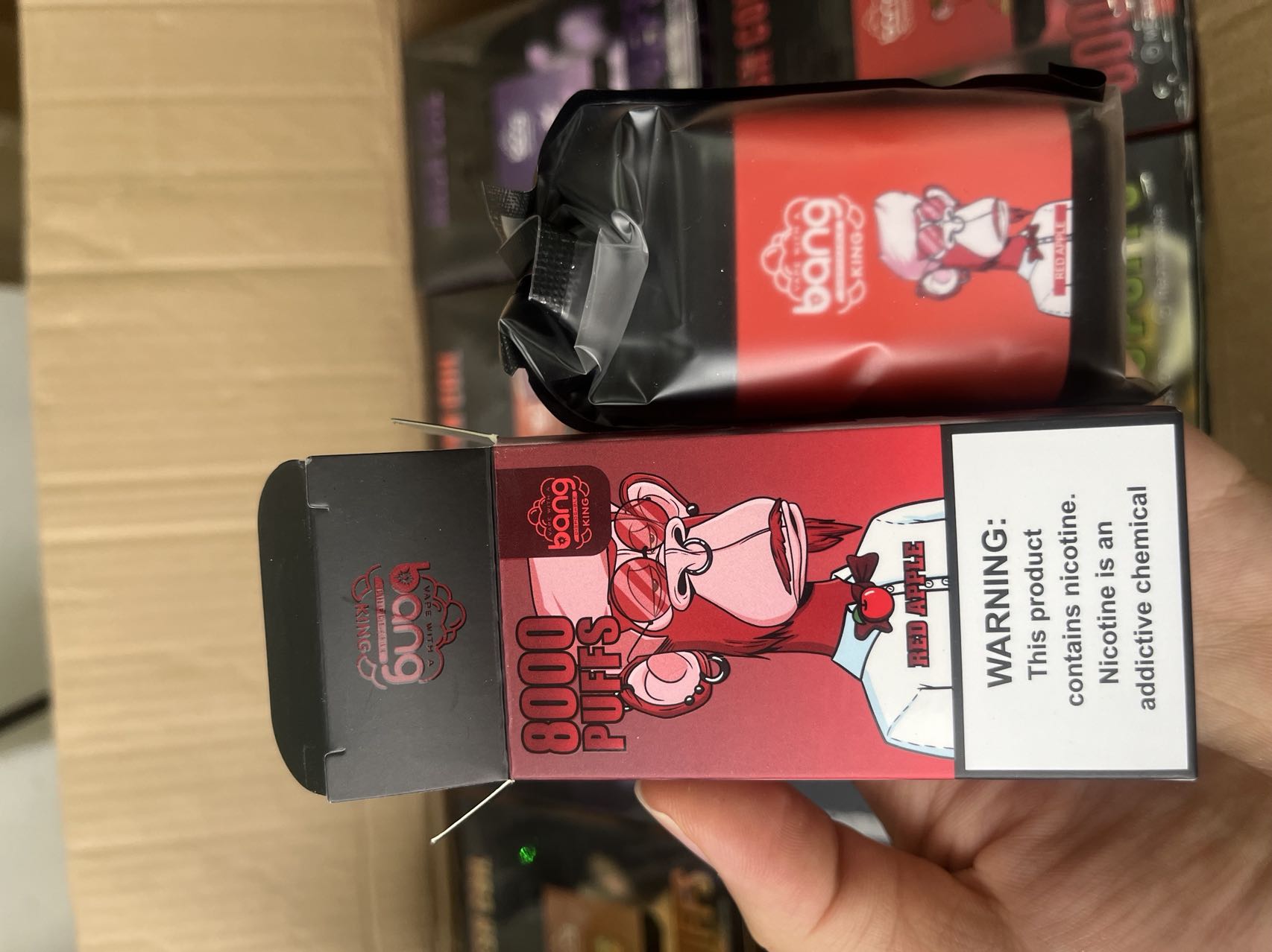 A hand holding a black and red vape device box labeled "Bang 8000 Puffs" with a cartoon ape and the flavor "Red Apple." A nicotine warning is visible on the box.