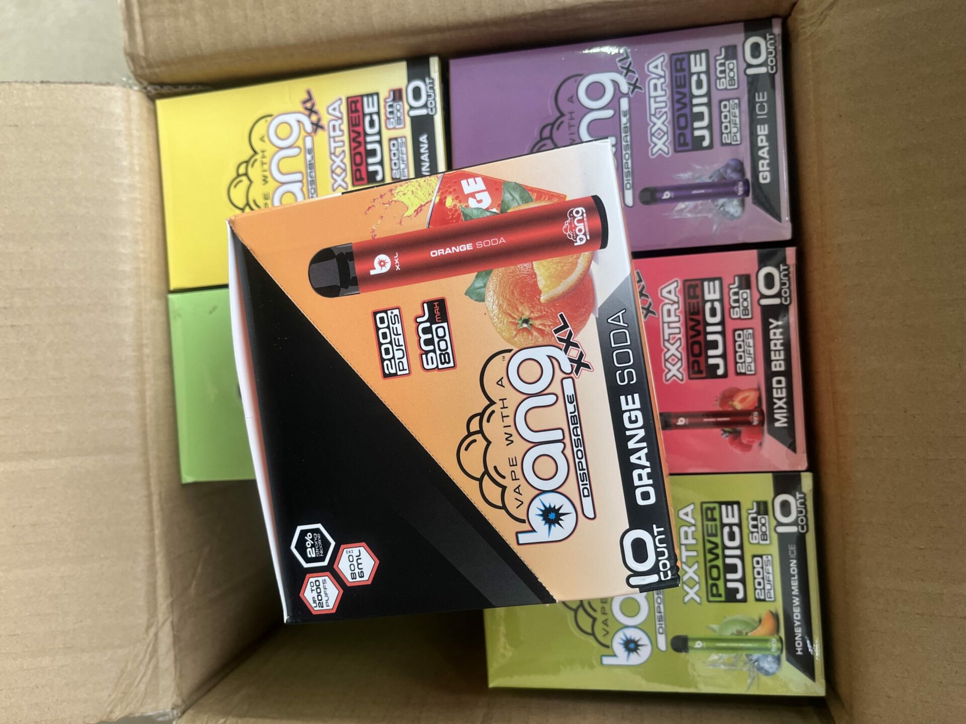 A box of assorted Bang XXL disposable vape pens, with visible flavors including Orange Soda, Grape Ice, Mixed Berry, and Honeydew Melon Ice.