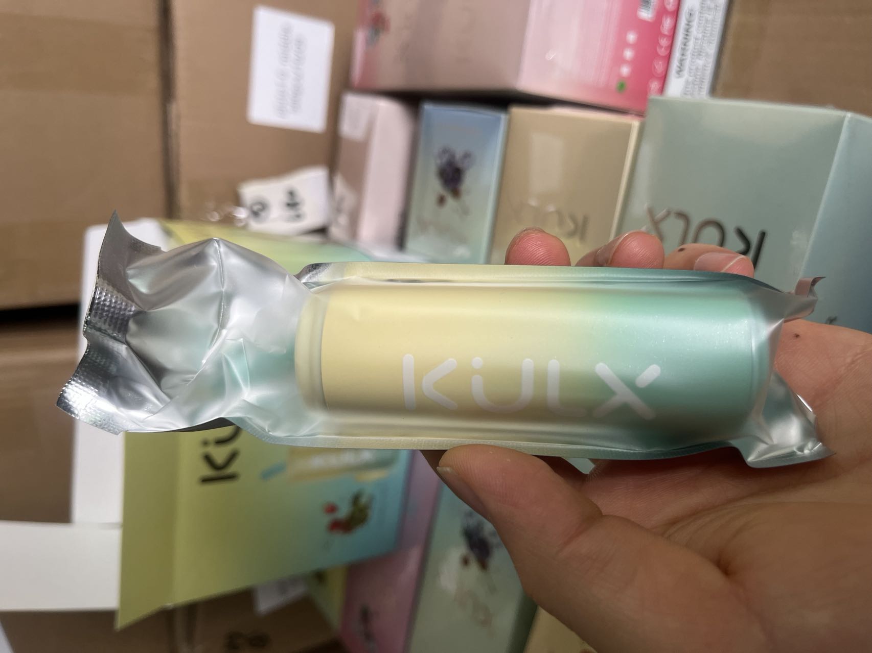 A hand holding a cylindrical, pastel-colored product with the brand name "KULX" printed on it, packaged in a transparent wrapper.