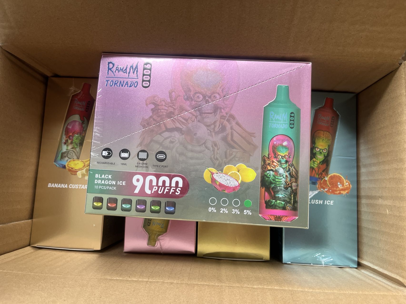 A cardboard box containing various flavors of RandM Tornado disposable vape devices, with the box for "Black Dragon Ice" flavor prominently displayed, showing 9,000 puffs and nicotine strength options.