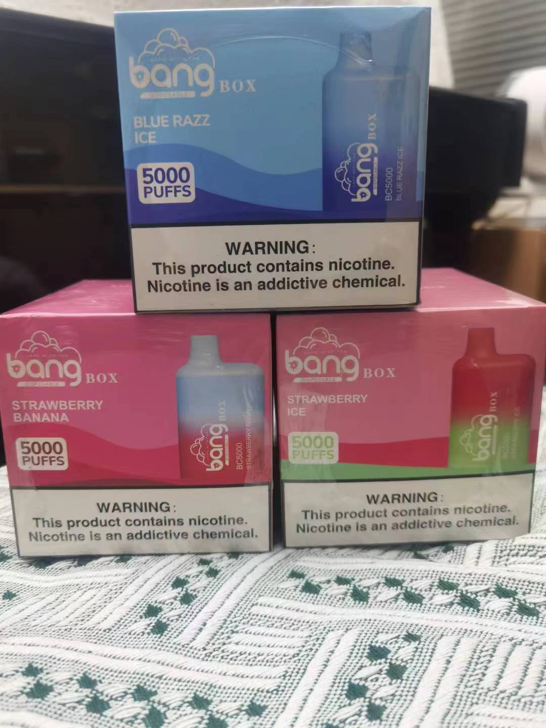 Three boxes of Bang disposable vapes in different flavors (Blue Razz Ice, Strawberry Banana, and Strawberry Ice) stacked on a patterned surface. Each box is labeled with "5000 PUFFS" and has a nicotine warning at the bottom.