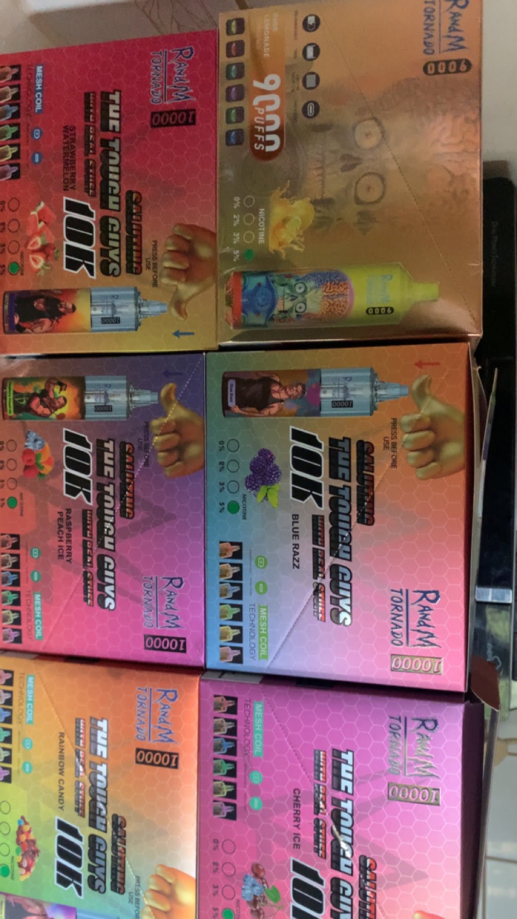 Boxes of disposable vape products labeled "RAWAM Tornado" and "The Tough Guys 10K," featuring different flavors such as strawberry watermelon, blue razz, and cherry ice, with images of the vape device and puff count details on the packaging.