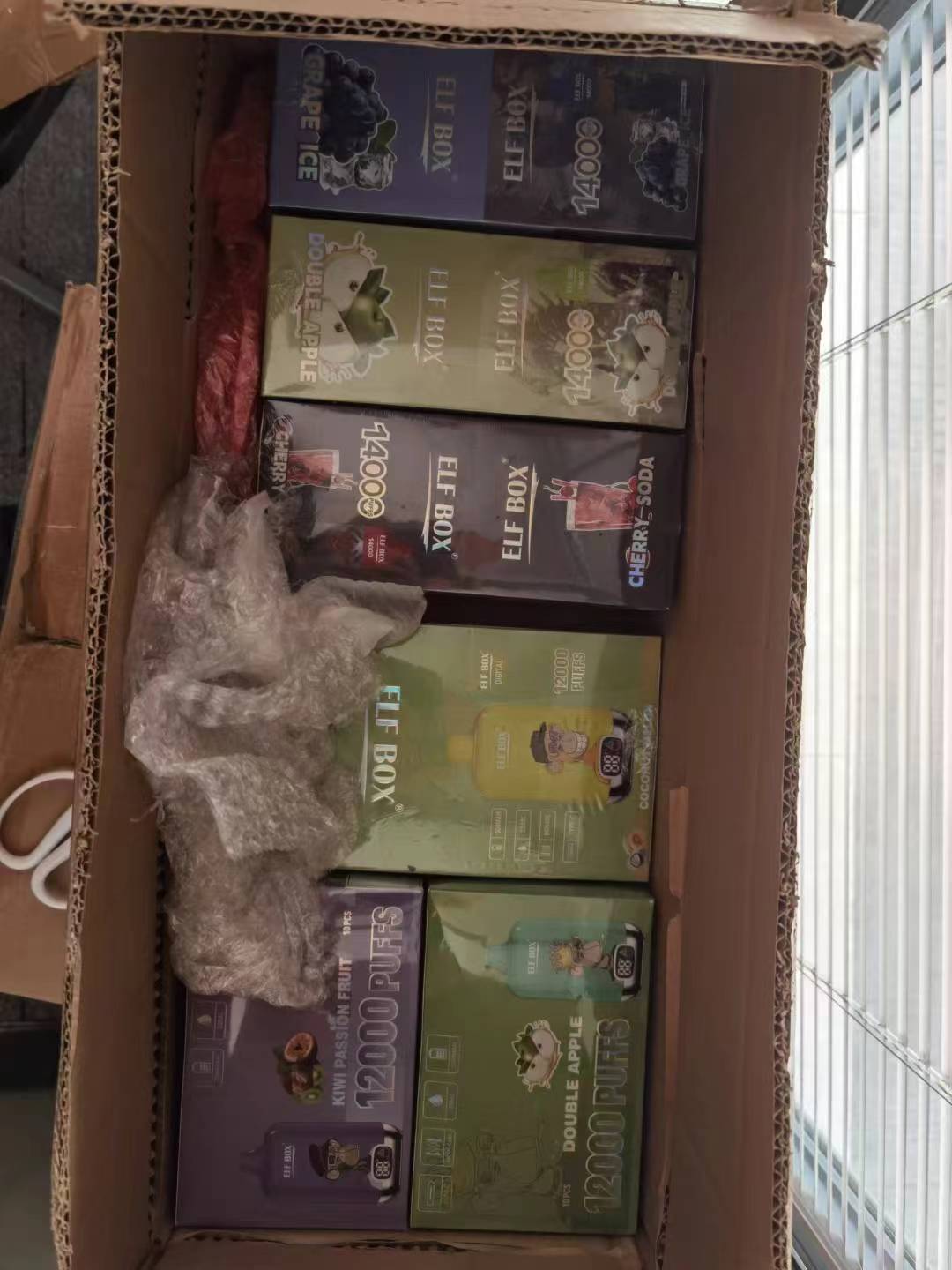 A box containing various Elf Box disposable vape packaging, including flavors like Grape Ice, Double Apple, and Cherry Soda.