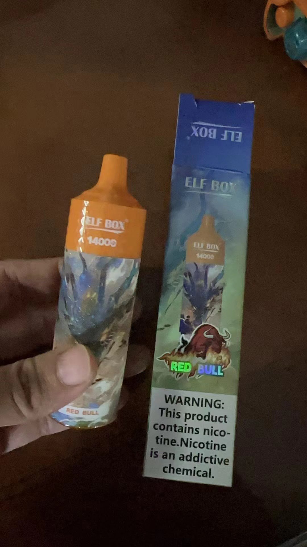 A hand holding an "Elf Box 14000" vape device with Red Bull flavor, alongside its matching box displaying the same branding and a nicotine warning.