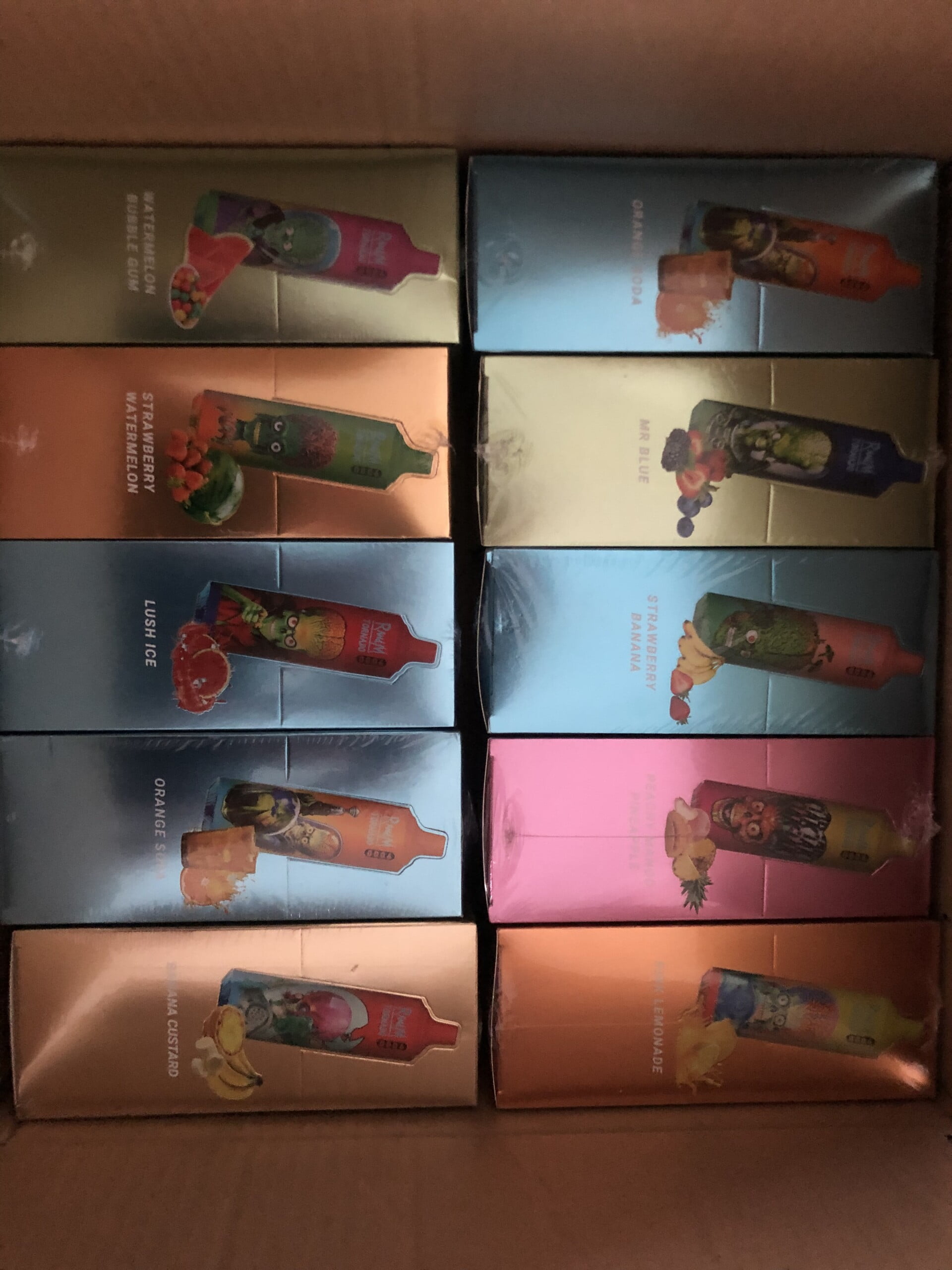 A box containing colorful vape packages, arranged in rows with different flavor labels such as "Watermelon Bubble Gum," "Strawberry Watermelon," and "Orange Soda."