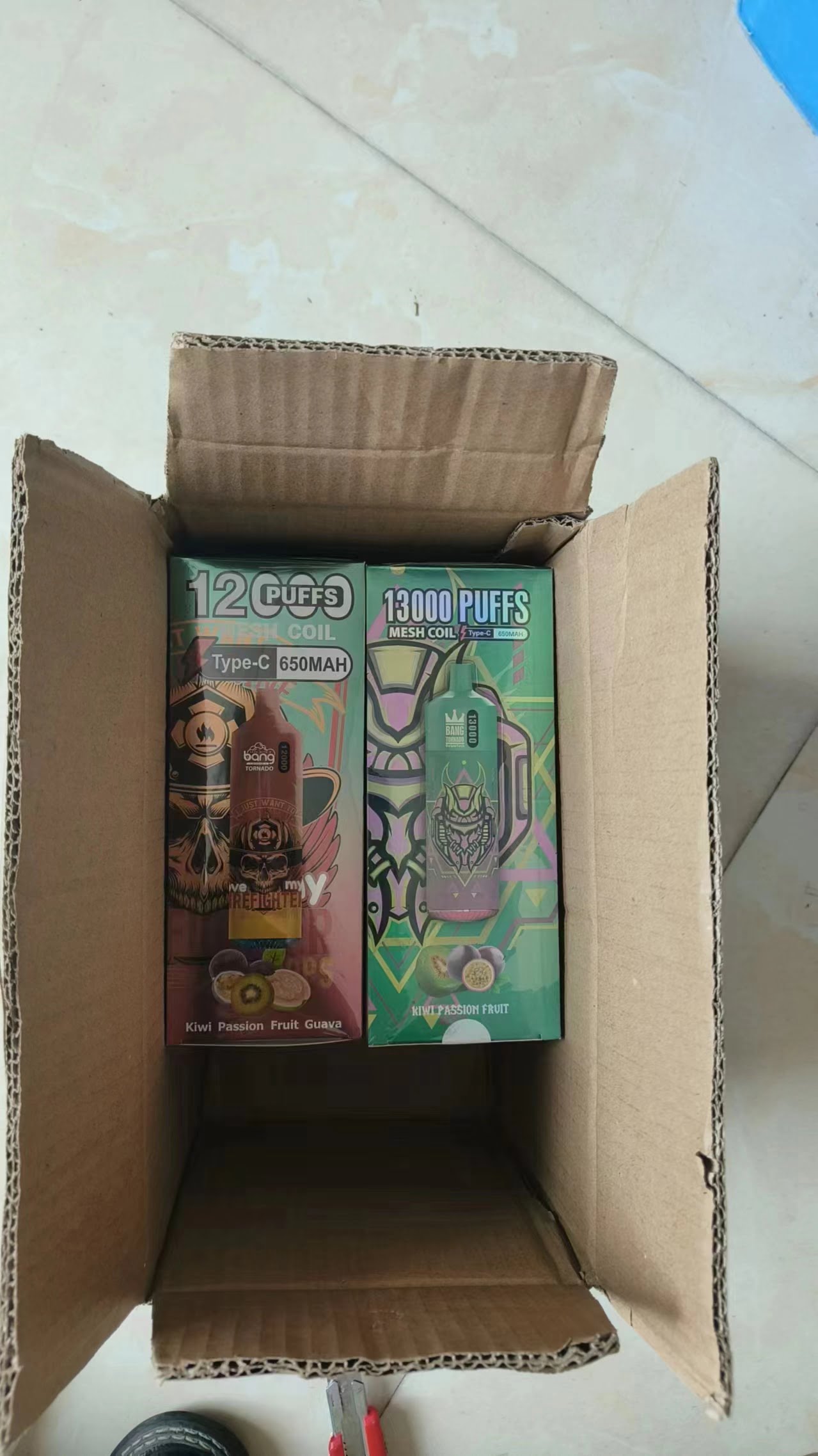 Two boxed electronic vaping devices inside an open cardboard box, labeled with "13000 PUFFS" and "12000 PUFFS," featuring Kiwi Passion Fruit and Guava flavors.