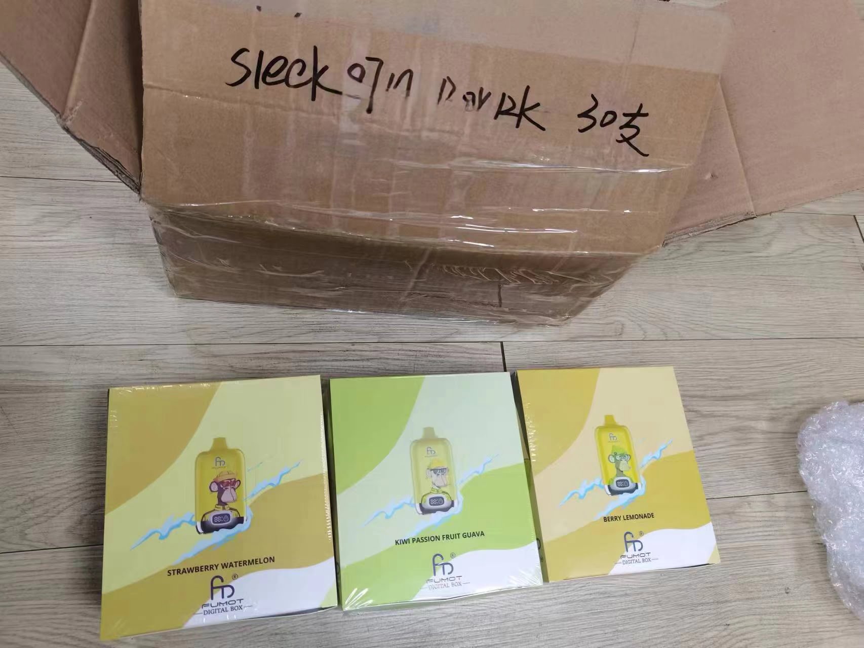 A cardboard box with black handwritten text on top, containing three packaged boxes labeled "STRAWBERRY WATERMELON," "KIWI PASSION FRUIT GUAVA," and "BERRY LEMONADE," each with a yellow and white design.