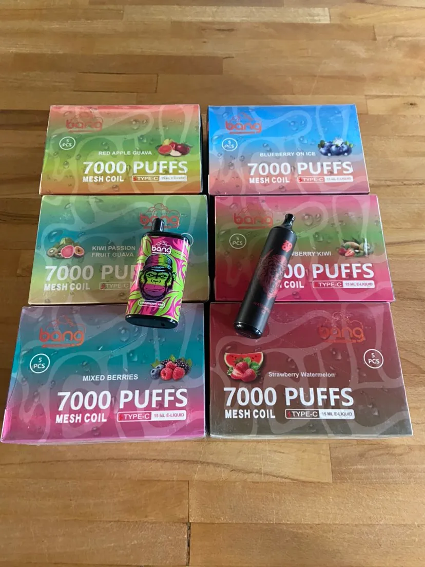 Boxes of Bang disposable vapes, each labeled "7000 PUFFS MESH COIL," arranged on a wooden surface. Two individual vape devices are placed in front of the boxes.