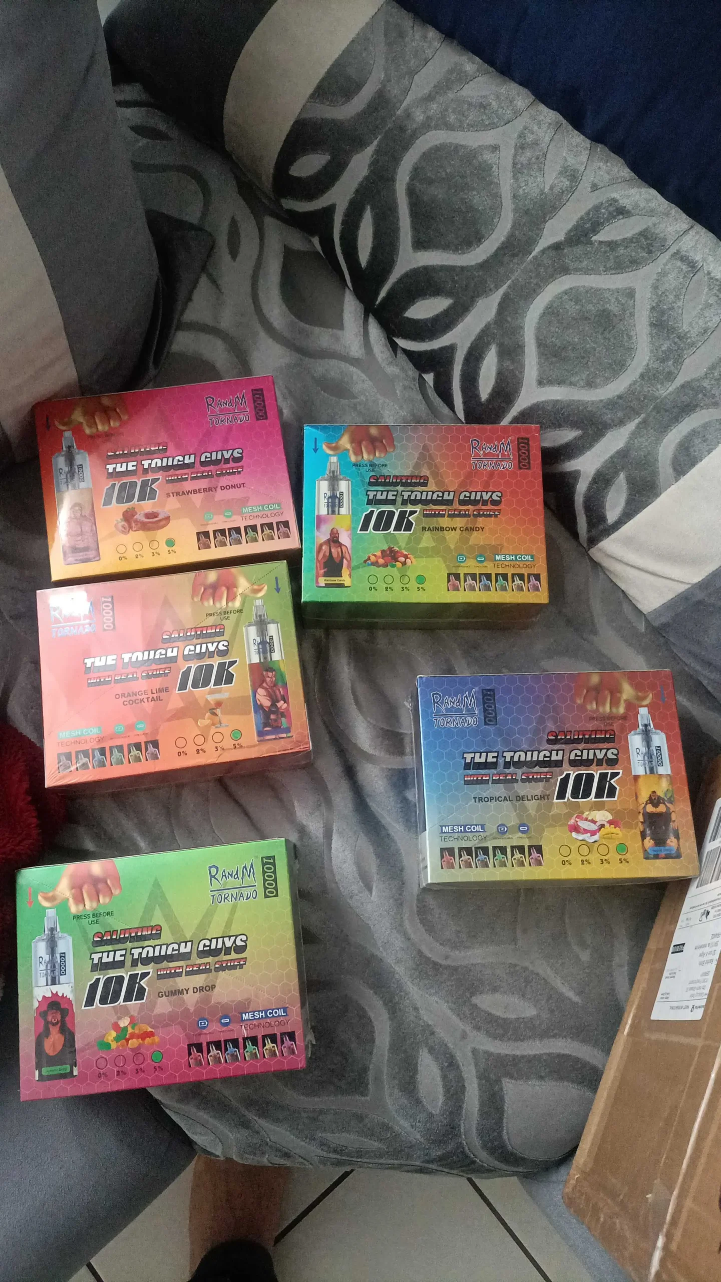 Five boxes of vaping devices with colorful packaging labeled "The Tough Guys 10K," featuring different flavors such as Strawberry Donut, Rainbow Candy, and Tropical Delight, displayed on a gray-patterned surface.