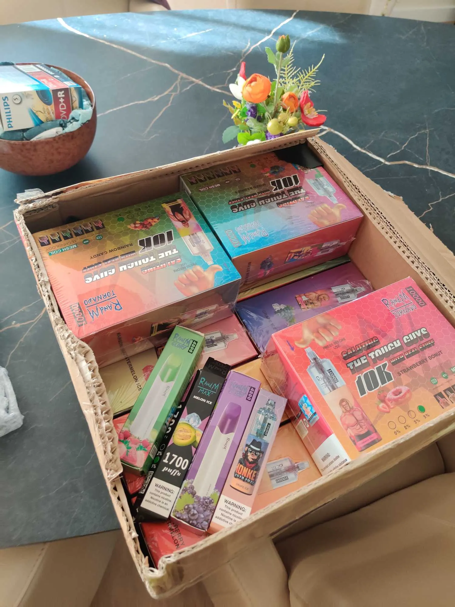 A cardboard box filled with various brightly colored disposable vape packages placed on a dark marble table next to a small flower arrangement and a bowl containing a Philips DVD+R spindle.