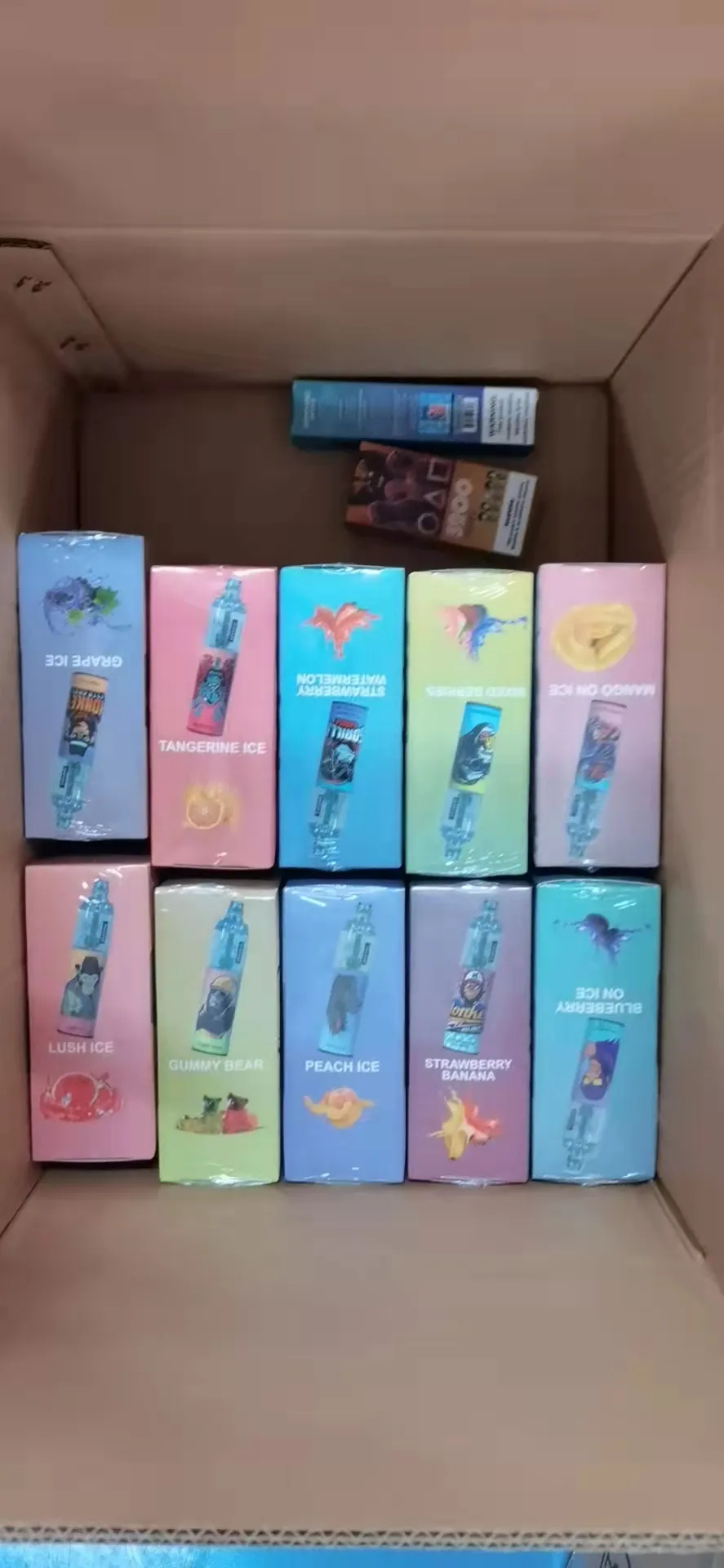 A cardboard box containing various boxed vape products, arranged neatly in two rows, with flavors like "Grape Ice," "Tangerine Ice," and "Strawberry Banana" visible.