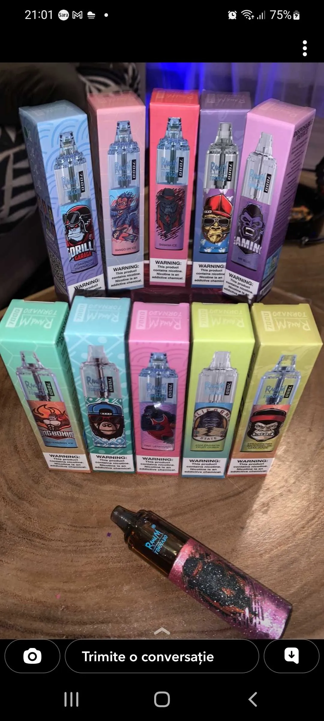 A variety of colorful vape product boxes arranged in two rows on a wooden surface, each featuring different graphic designs and flavor names. One vape device lies in front of the boxes.