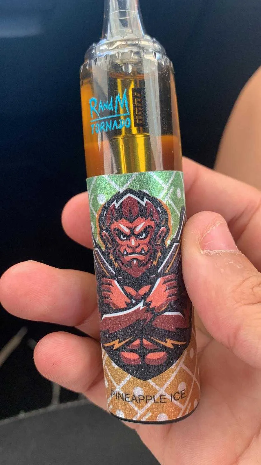 A hand holding a vape device labeled "RandM Tornado" with an illustration of a fierce-looking creature and the text "Pineapple Ice."