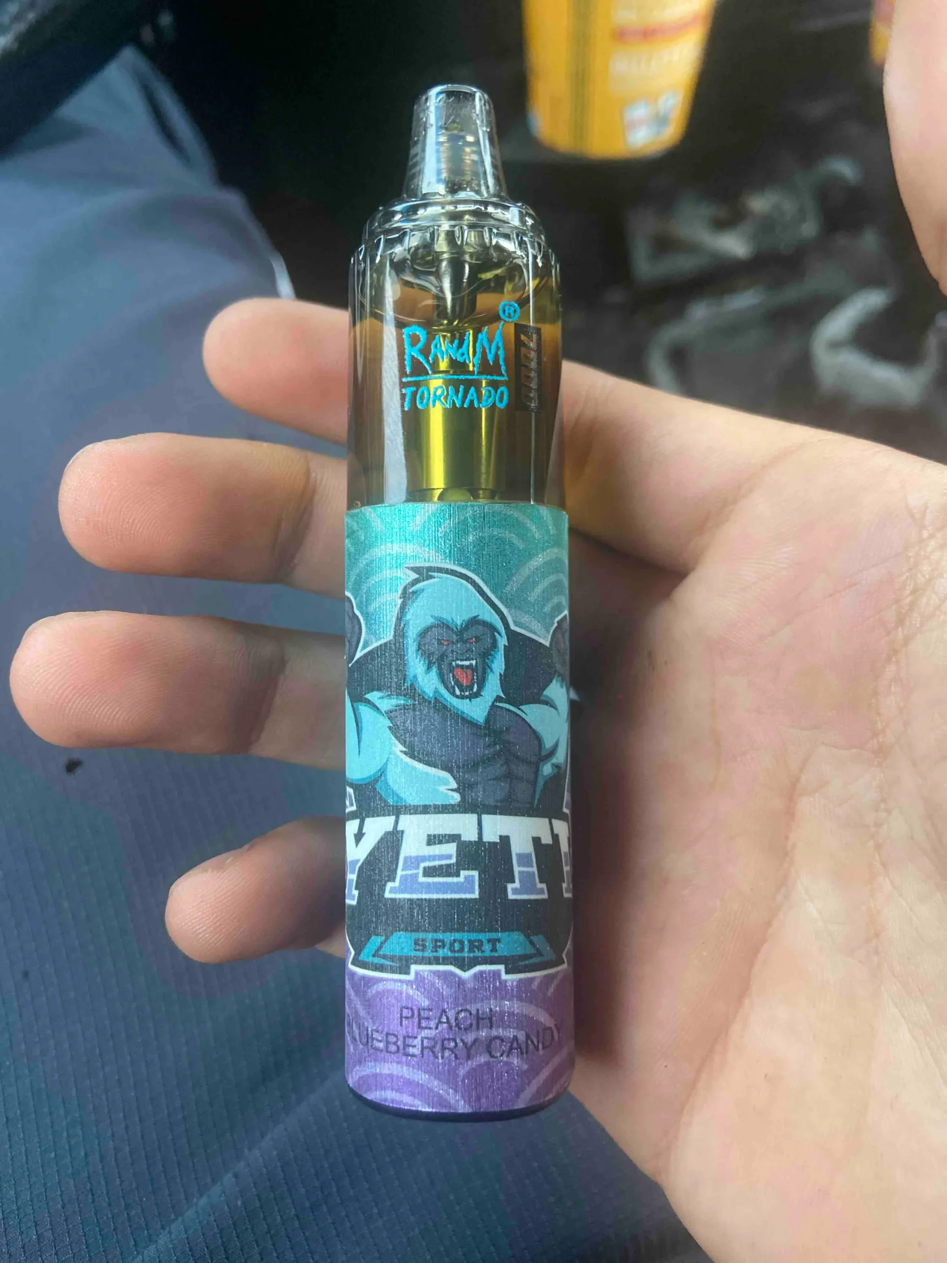 A person holding a disposable vape pen labeled "RAWM Tornado Yeti Sport" with "Peach Blueberry Candy" flavor.