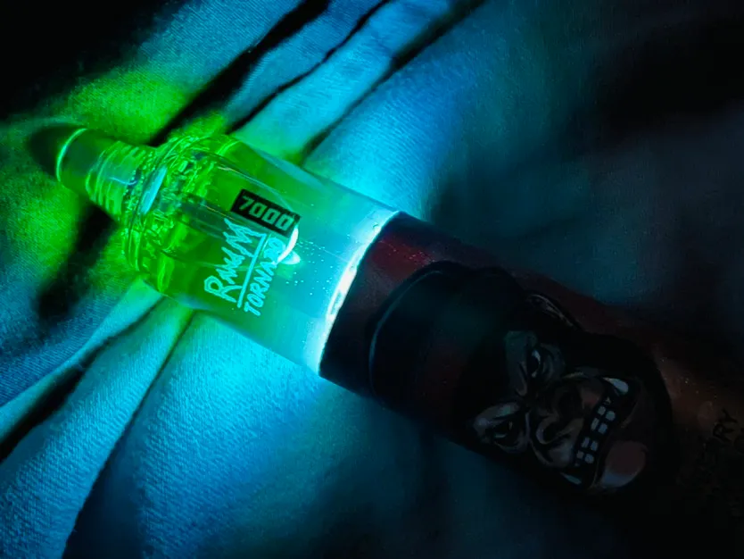 A green and blue glowing object, resembling a bottle with a graphic of a gorilla face, lying on a textured fabric background.