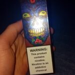 A hand holding a colorful vape box with a warning label stating, "This product contains nicotine. Nicotine is an addictive chemical."