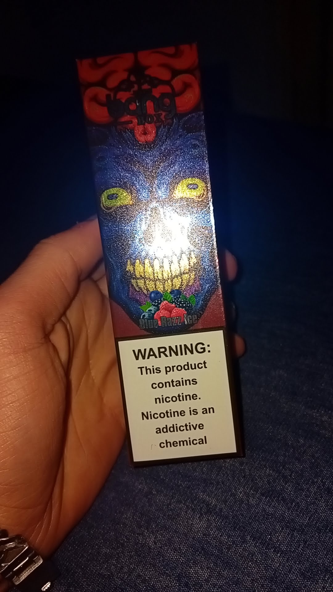 A hand holding a colorful vape box with a warning label stating, "This product contains nicotine. Nicotine is an addictive chemical."