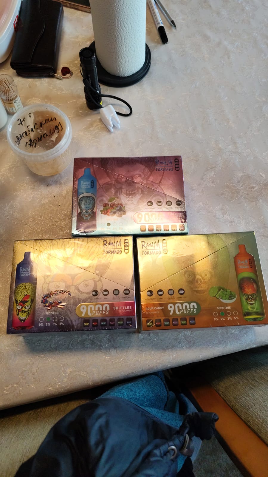 Three boxes of "Ranih Tornado 9000 Puffs" disposable vapes with different flavors on a table, along with a container of food, a paper towel roll, a flashlight, and other small items.