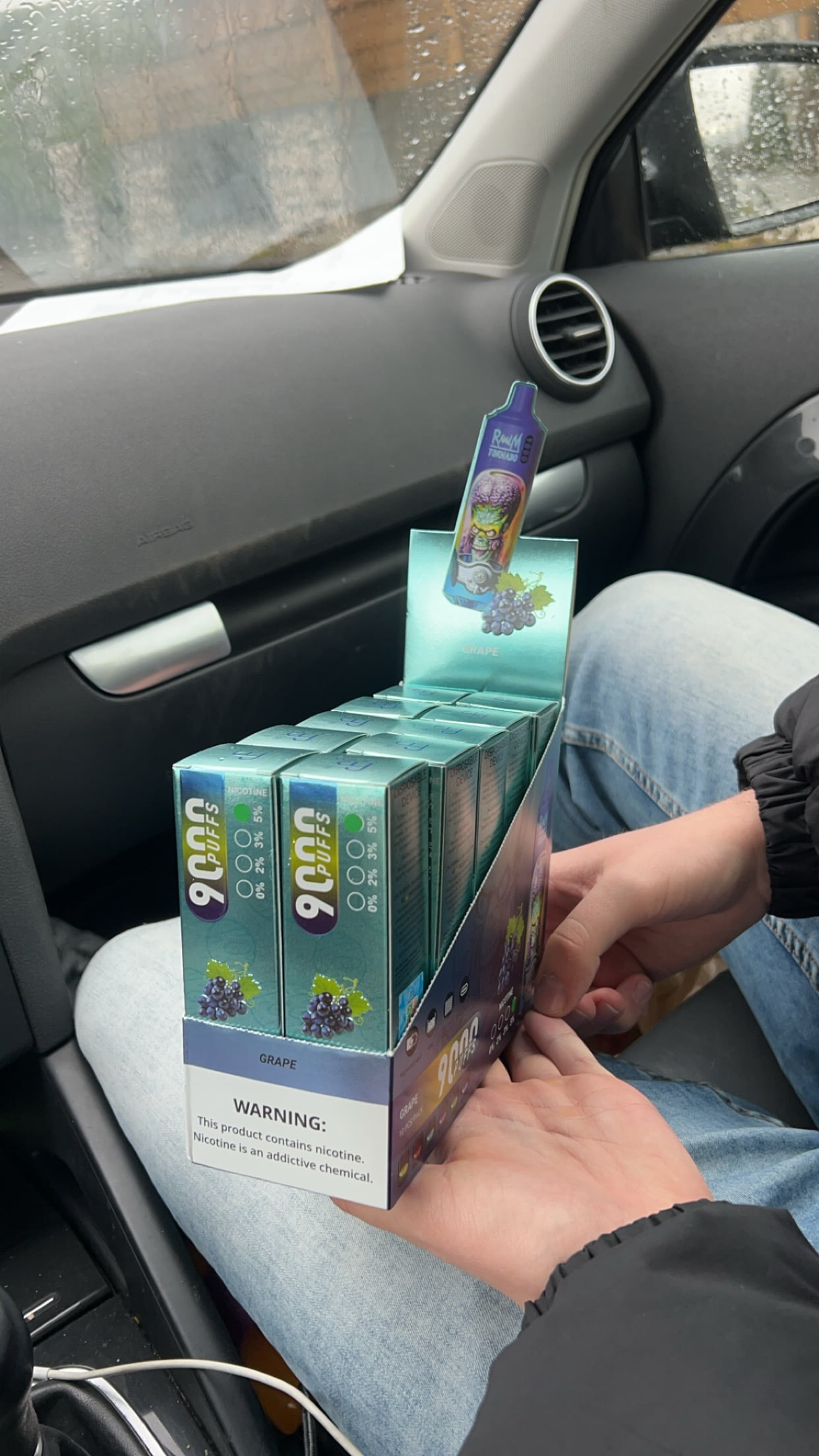 A person holding a box of disposable vape pens labeled "9000 puffs," with grape flavor and a warning about nicotine addiction.