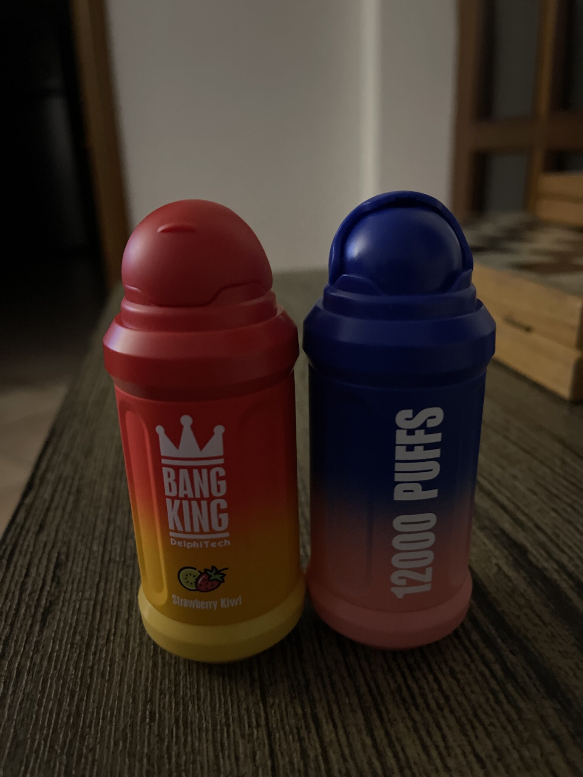 Two colorful disposable vape devices on a table: one red and yellow labeled "BANG KING" with a strawberry kiwi flavor, the other blue and pink labeled "12000 PUFFS."