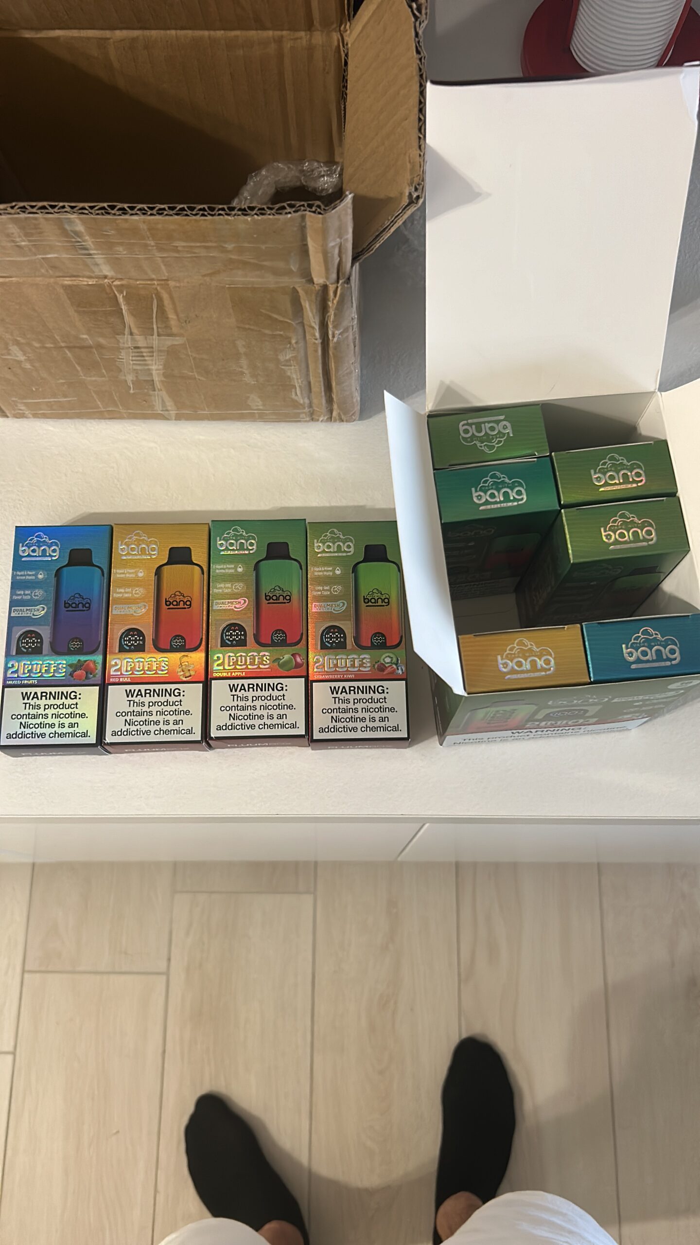 A row of Bang disposable vape packages in various flavors arranged on a white countertop next to an open cardboard box.