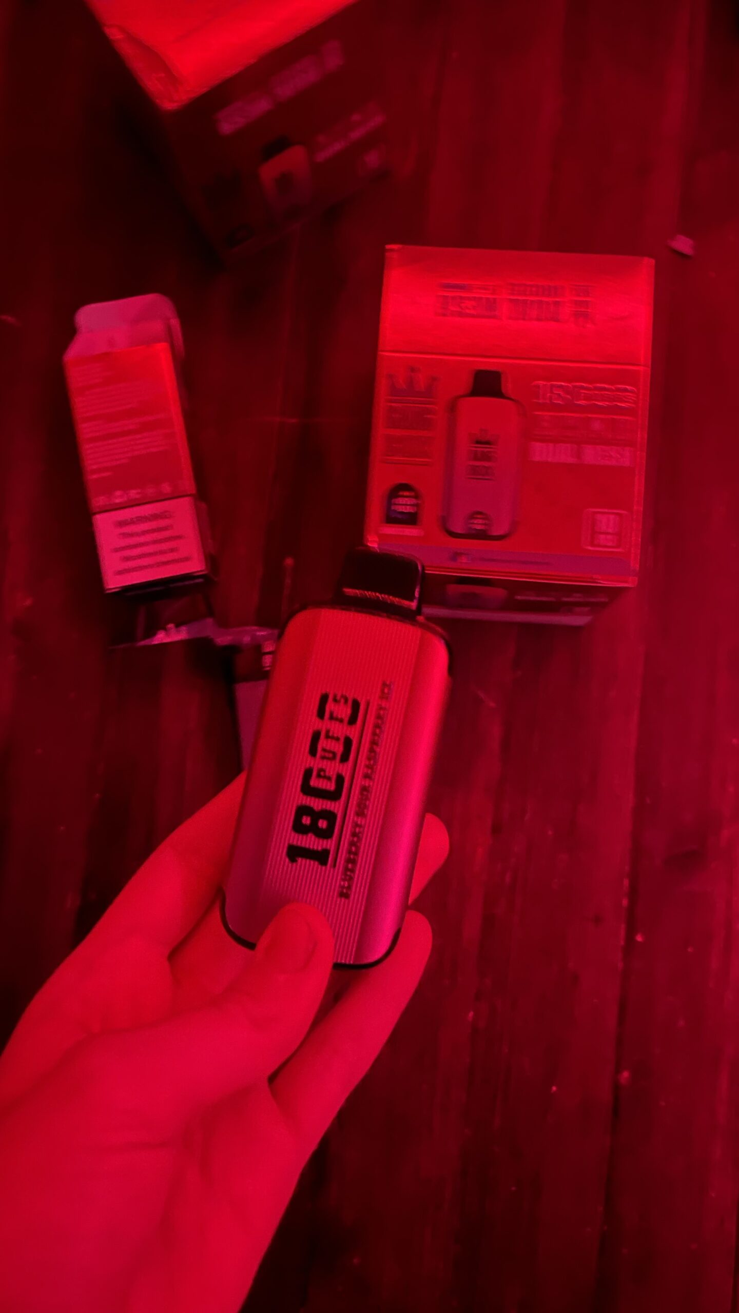 A hand holding a vape device labeled "1800 puffs" in a red-lit environment, with packaging and other vape items visible in the background.